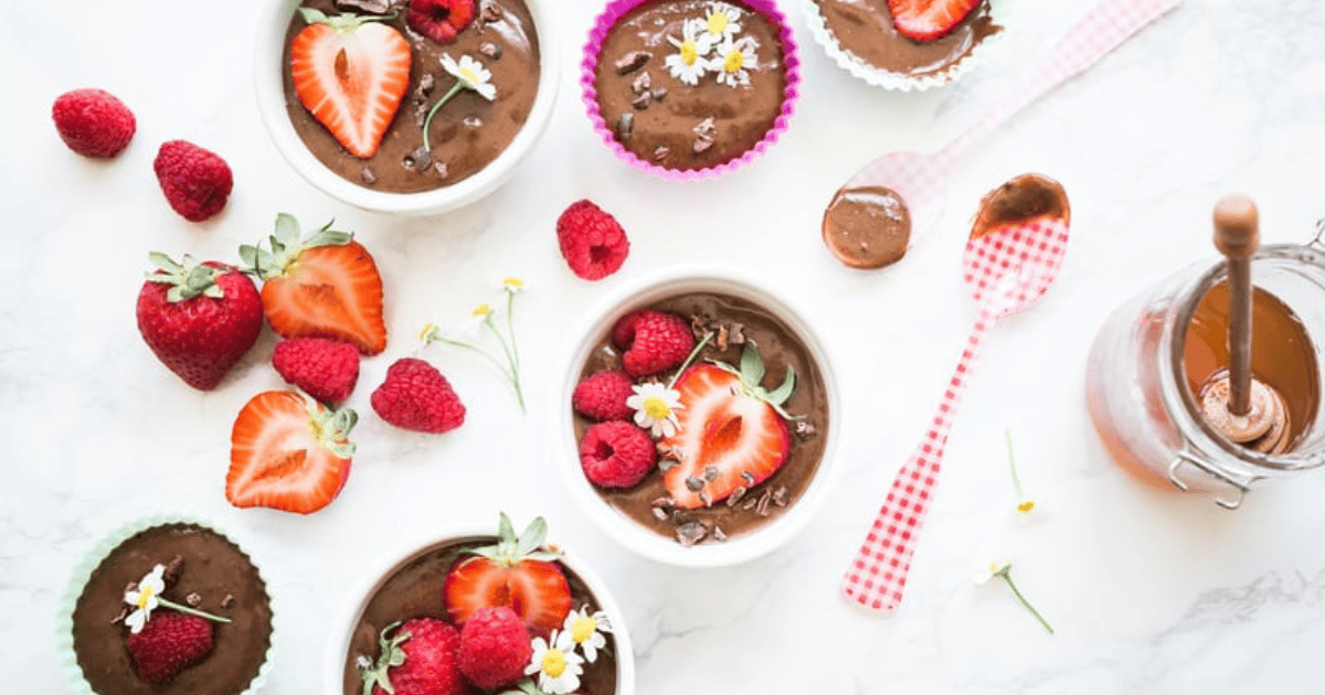 Bowls of chocolate mousse topped with strawberries, raspberries, cacao nibs, and small white flowers tempt anyone who loves dessert—making it easy to forget moderation and indulge in a moment of binge eating. Fresh berries and a jar of chocolate sauce are on the white surface, with a spoon and checkered napkin beside them.