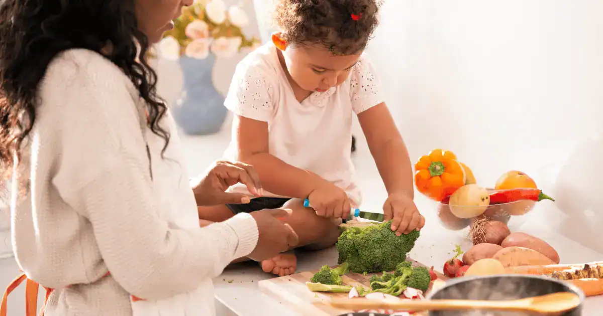 A young child sits on a kitchen counter cutting broccoli with help from an adult, taking the first steps toward raising healthy eaters. Various vegetables are spread on the counter around them.